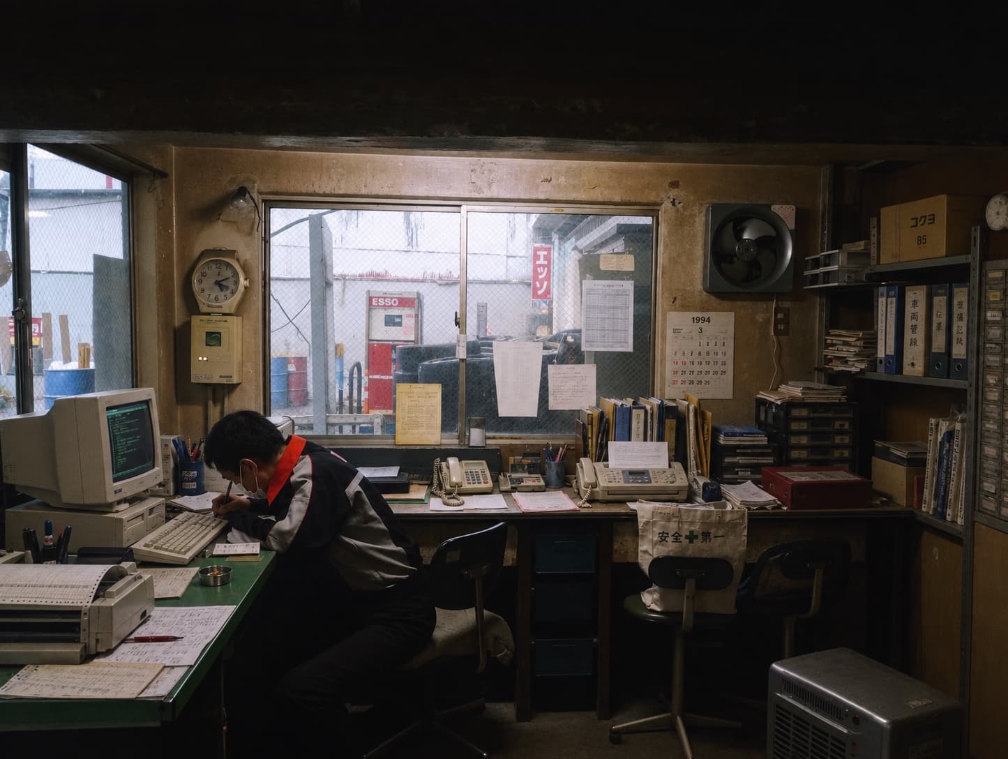 A 1990s back office: a person at a desktop computer beside a phone, a fax machine and stacks of paper.
